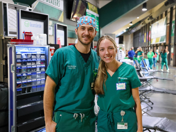 A male and femal in green scrubs smiling for a photo in front of dental equipment and chairs set up in the UAB Bartow Arena and volunteers walking in the background